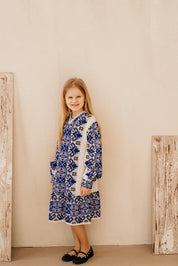 Young girl wearing a traditional Ukrainian style knitted embroidered dress in blue and white, standing with a smile against a beige backdrop.