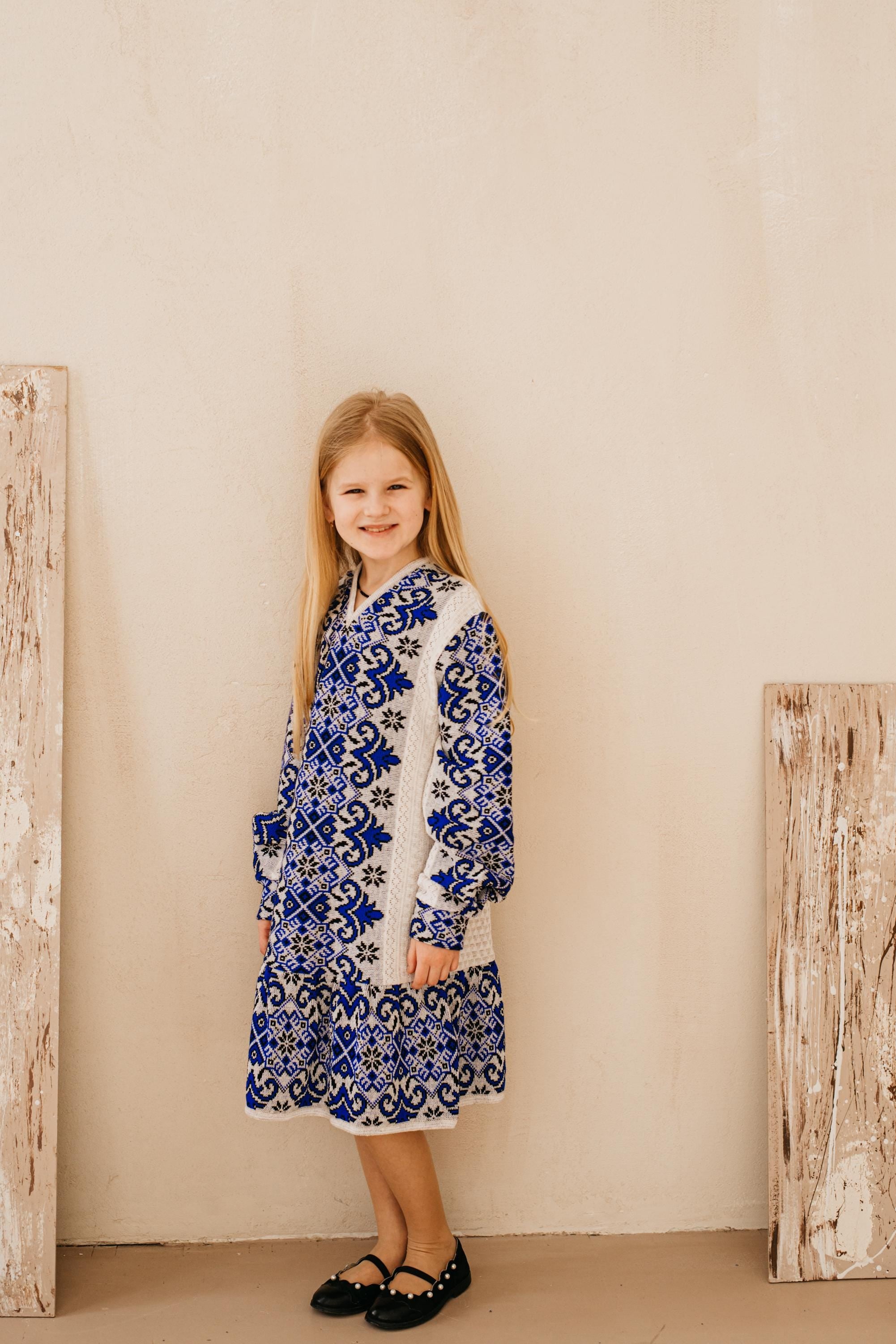 Young girl wearing a traditional Ukrainian style knitted embroidered dress in blue and white, standing with a smile against a beige backdrop.