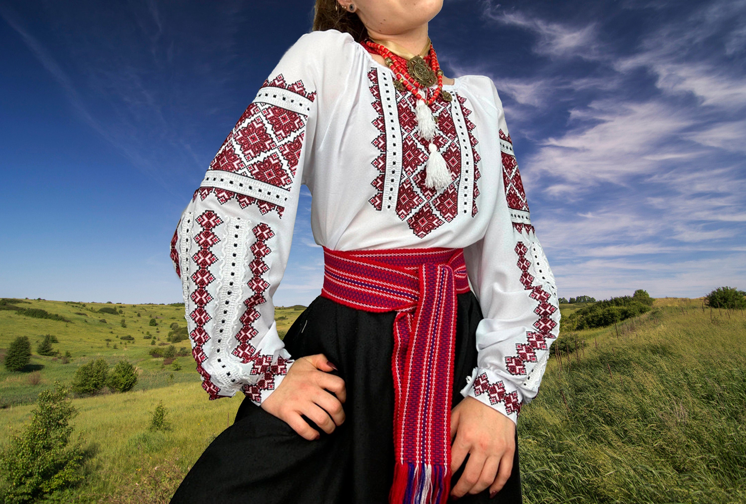 Woman wearing Ukrainian embroidered Vyshyvanka blouse with red and black cross-stitch design, standing in a scenic field.