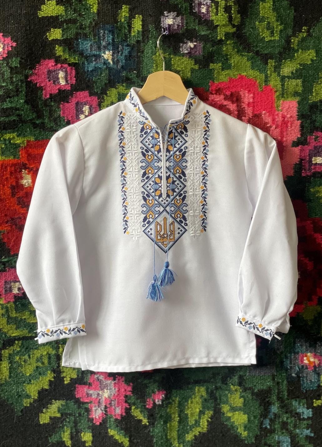 Ukrainian boy vyshyvanka shirt with Tryzub embroidery, white sorochka with blue and yellow design on floral background.