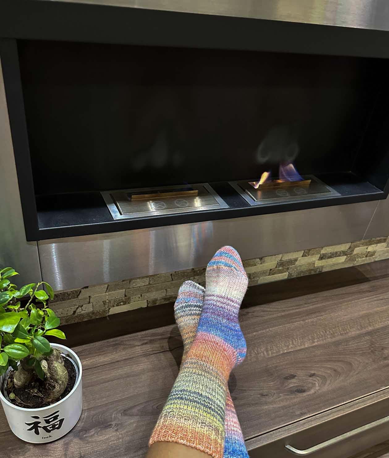 Cozy feet in colorful wool socks relaxing by a modern fireplace with a potted plant, showcasing warmth and comfort.