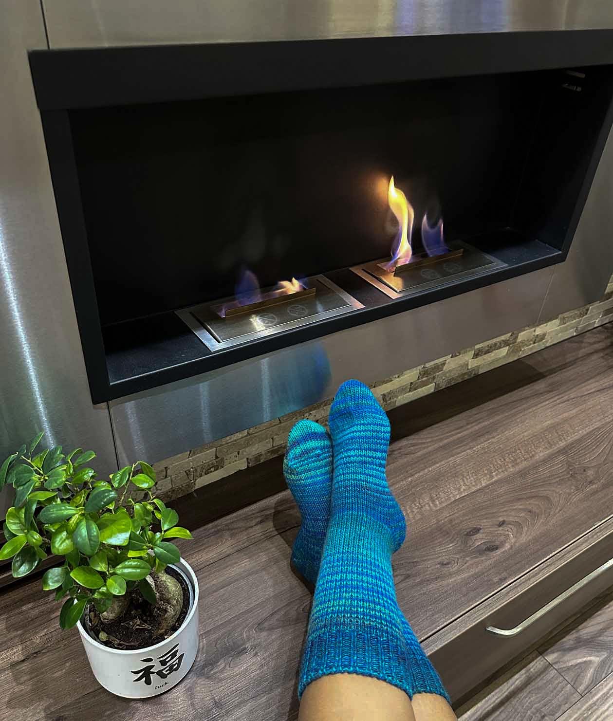 Person wearing blue wool socks by a cozy fireplace, next to a potted plant, staying warm during winter indoors.