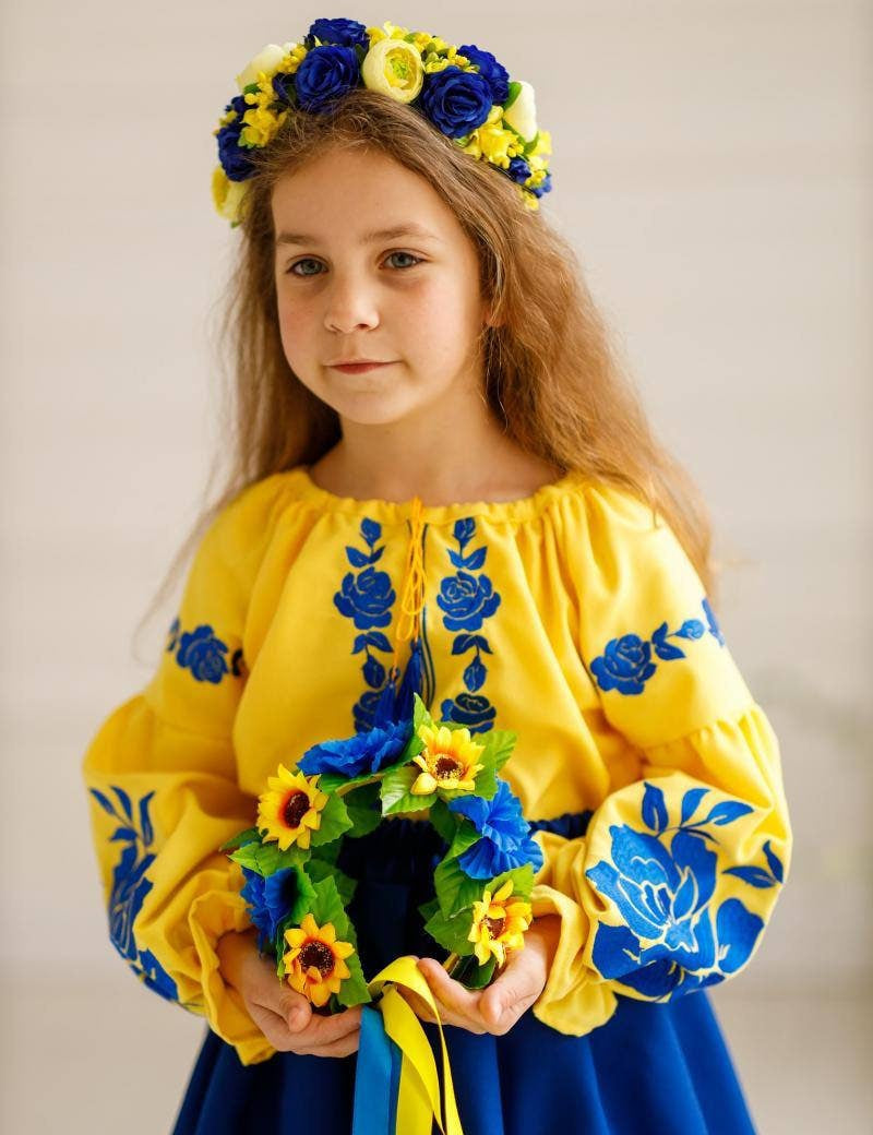 Girl wearing a Ukrainian embroidered blouse and skirt set in blue and yellow, holding a floral wreath, traditional outfit for cultural events.