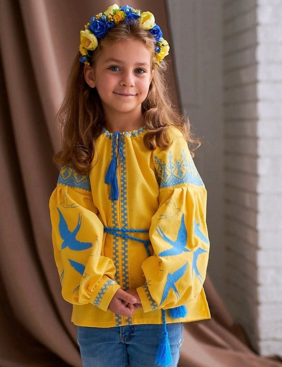 Girl in yellow Ukrainian embroidered blouse with blue swallow motif, wearing a blue and yellow flower crown.