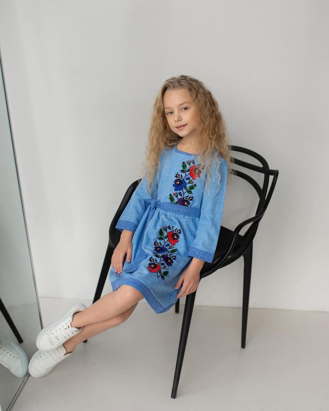 Girl in a blue Ukrainian embroidered dress with floral patterns sitting on a chair, showcasing traditional Slavic style.