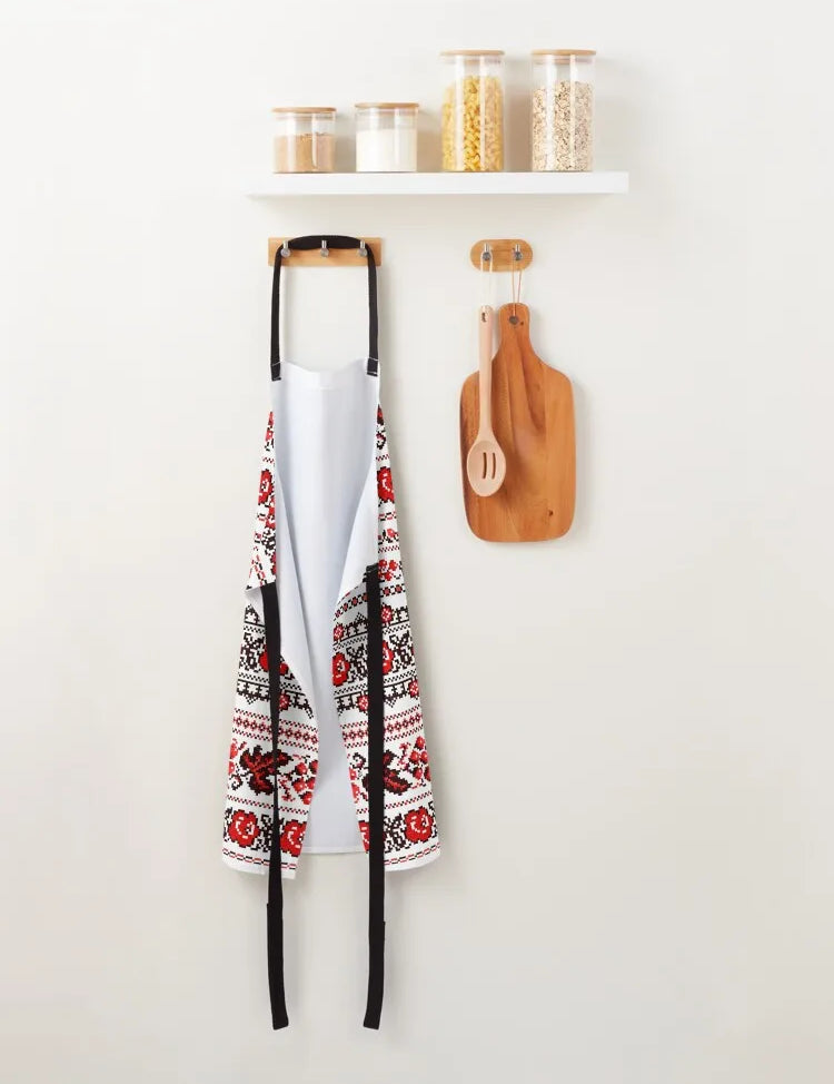 Ukrainian embroidered apron with red and black pattern hanging in a kitchen setting with culinary tools and spices on a shelf.