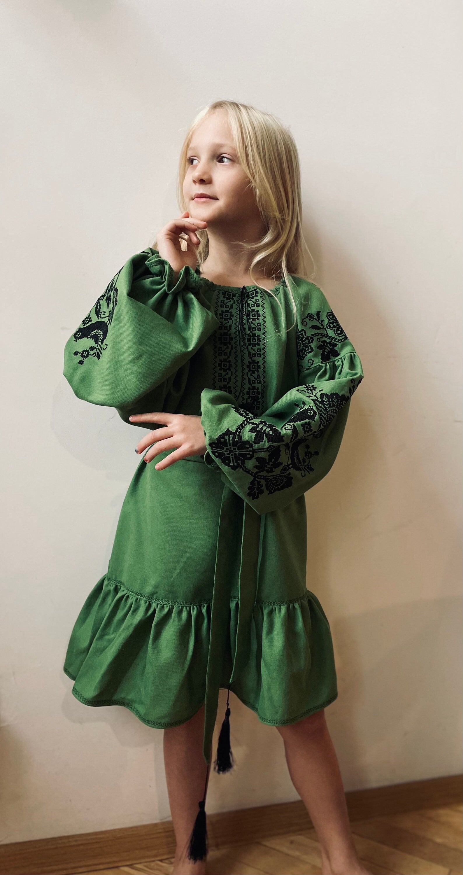 Girl wearing green embroidered Vyshyvanka dress in Ukrainian traditional style with black cross-stitch details, standing indoors.