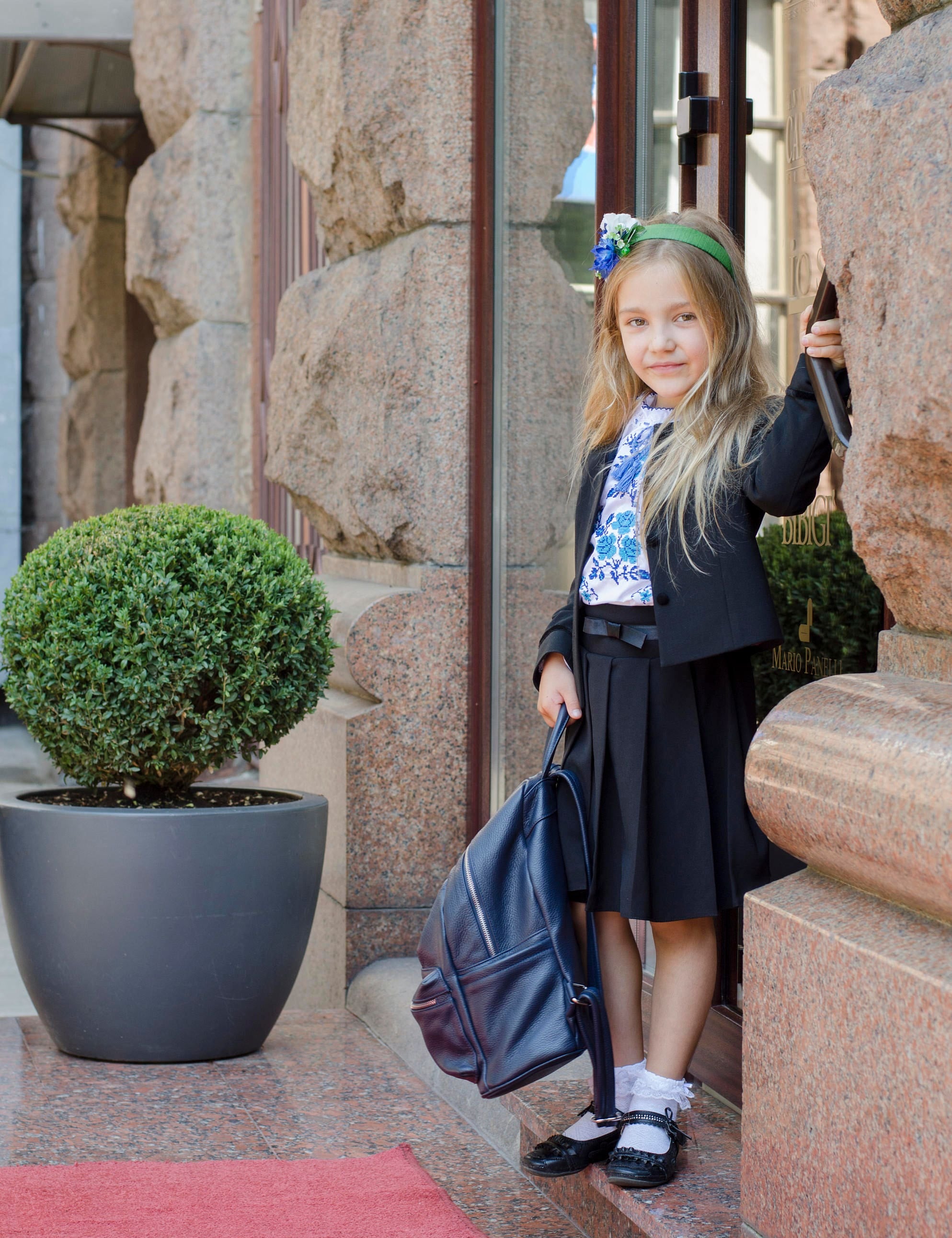 Young girl wearing a traditional Ukrainian embroidered shirt and black skirt, standing with a backpack, ready for a school event.
