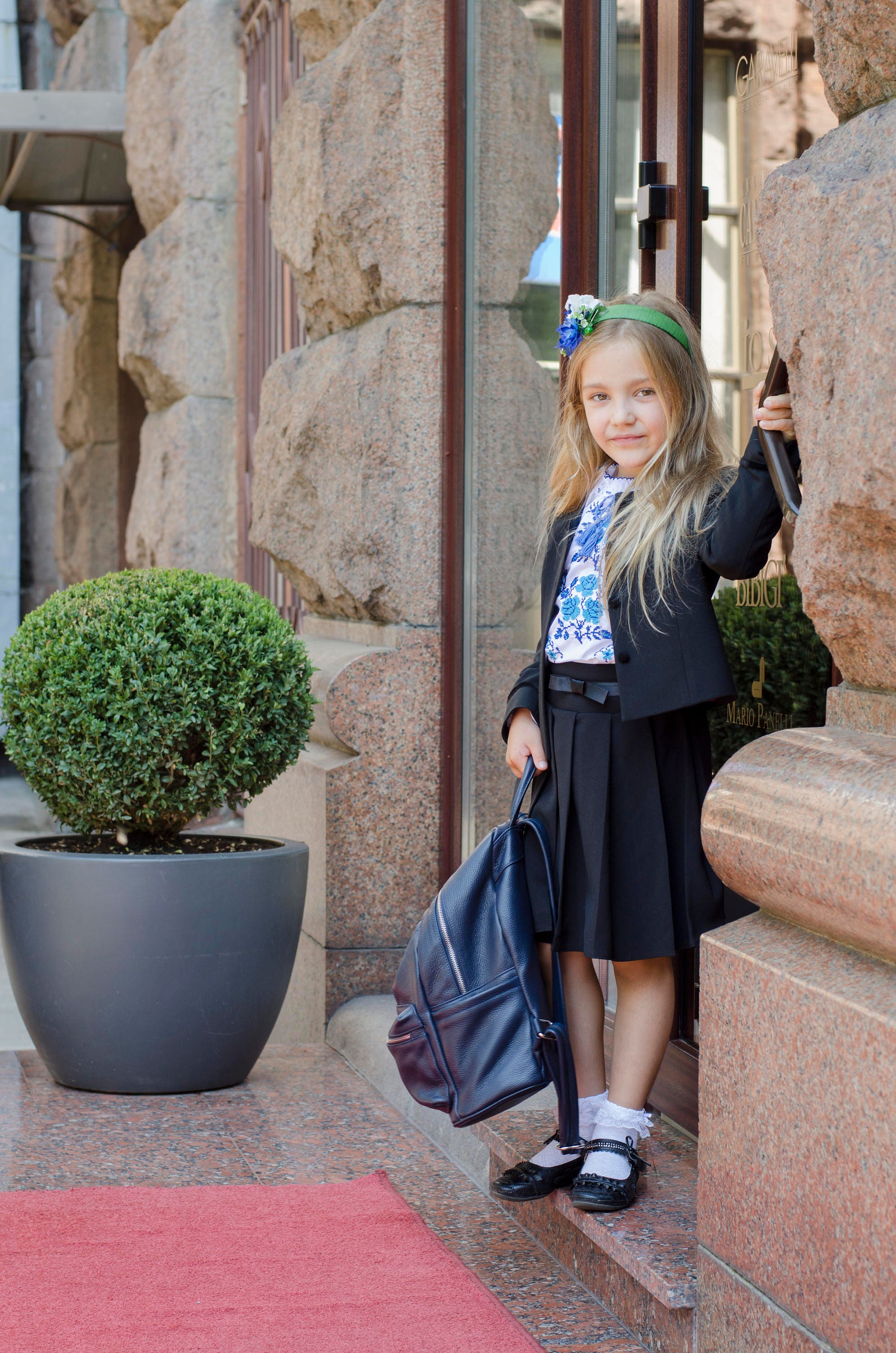 Young girl wearing a traditional Ukrainian embroidered shirt and black skirt, standing with a backpack, ready for a school event.