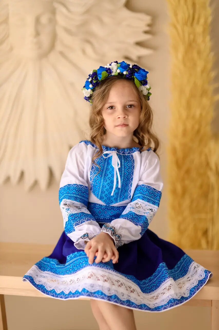 Girl wearing Ukrainian Hutsul blue folk costume with lace, seated with floral headband, traditional cultural attire for events.