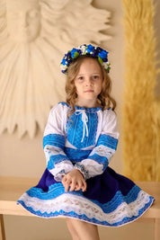 Girl wearing Ukrainian Hutsul blue folk costume with lace, seated with floral headband, traditional cultural attire for events.