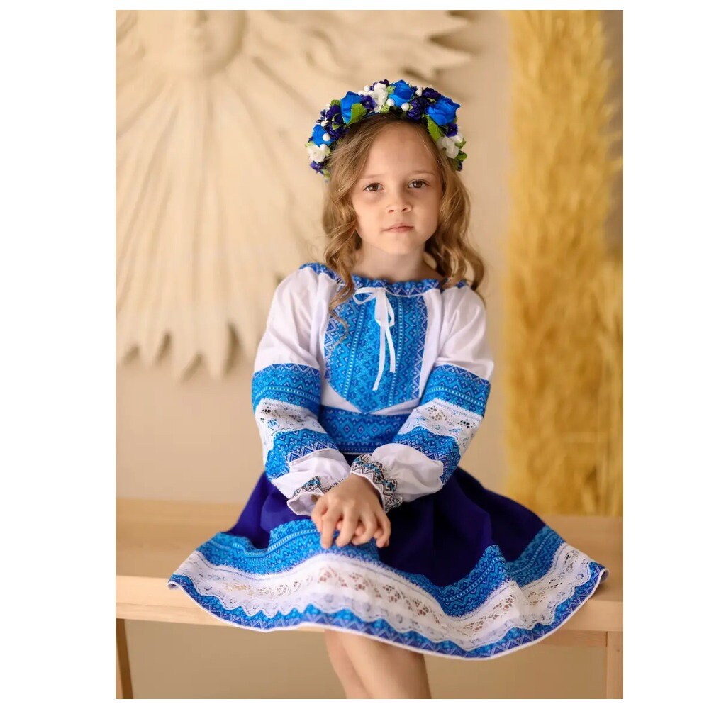 Girl in traditional Ukrainian Hutsul folk costume with blue woven blouse and skirt, wearing a floral headband, sitting on a bench.