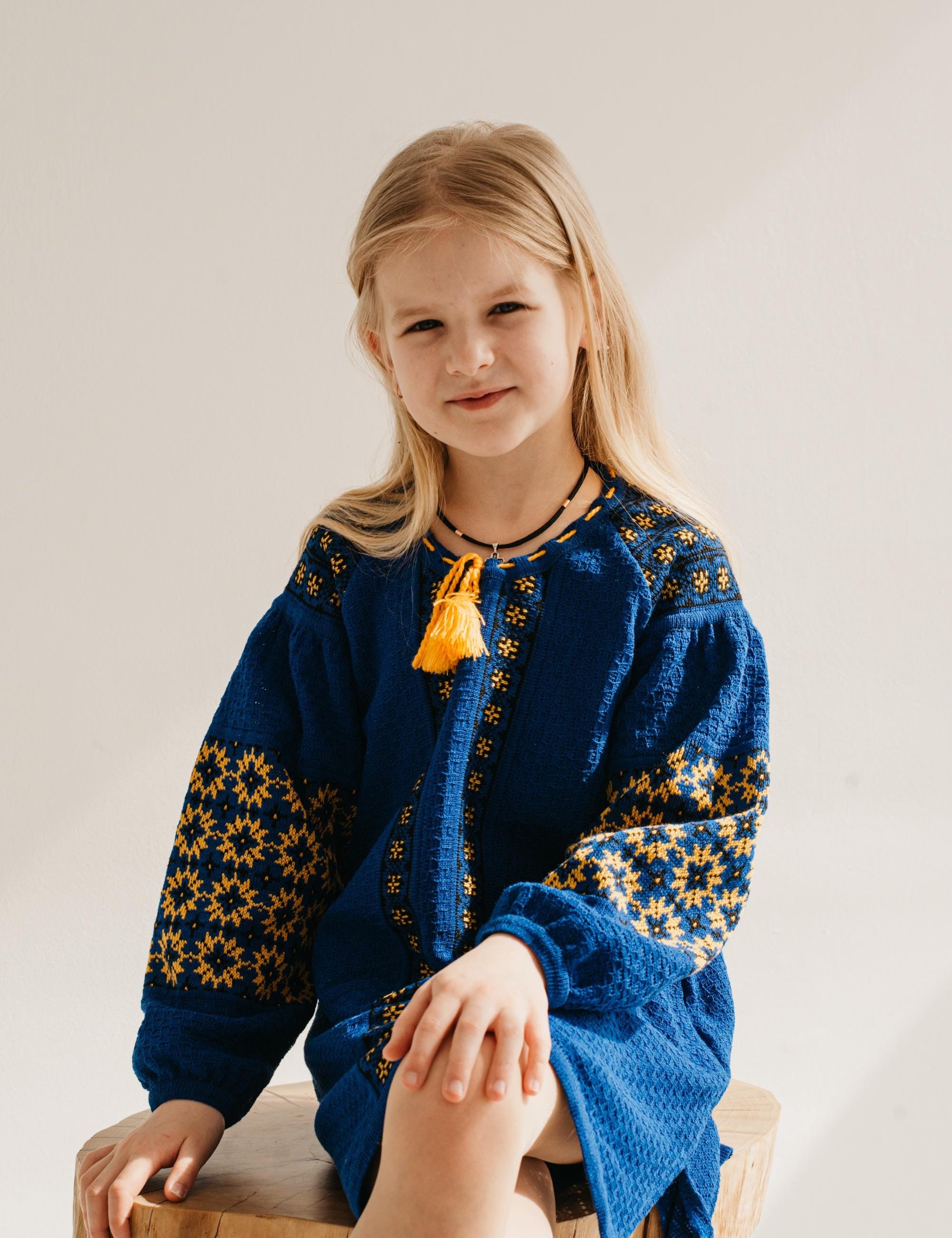 Young girl wearing a Ukrainian knitted dress with traditional embroidery, sitting on a wooden stool, embodying cultural style.