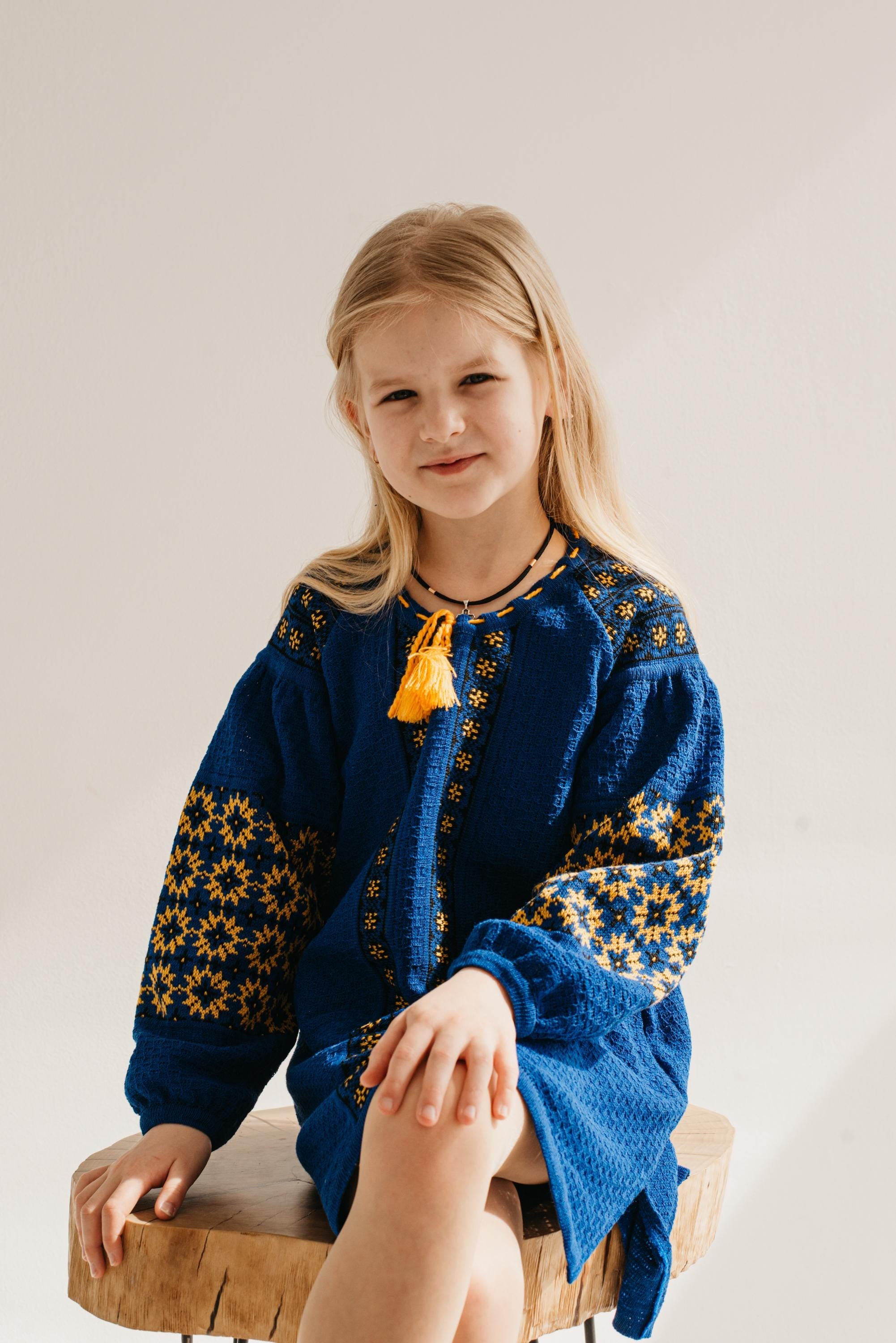 Young girl wearing a Ukrainian knitted dress with traditional embroidery, sitting on a wooden stool, embodying cultural style.