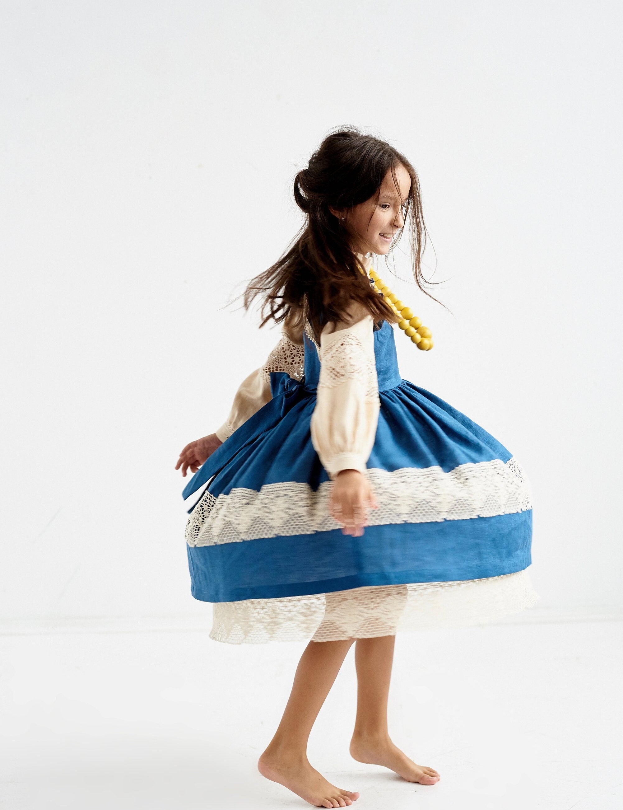 Child wearing a blue Ukrainian linen dress with lace trim and puff sleeves, spinning around playfully.