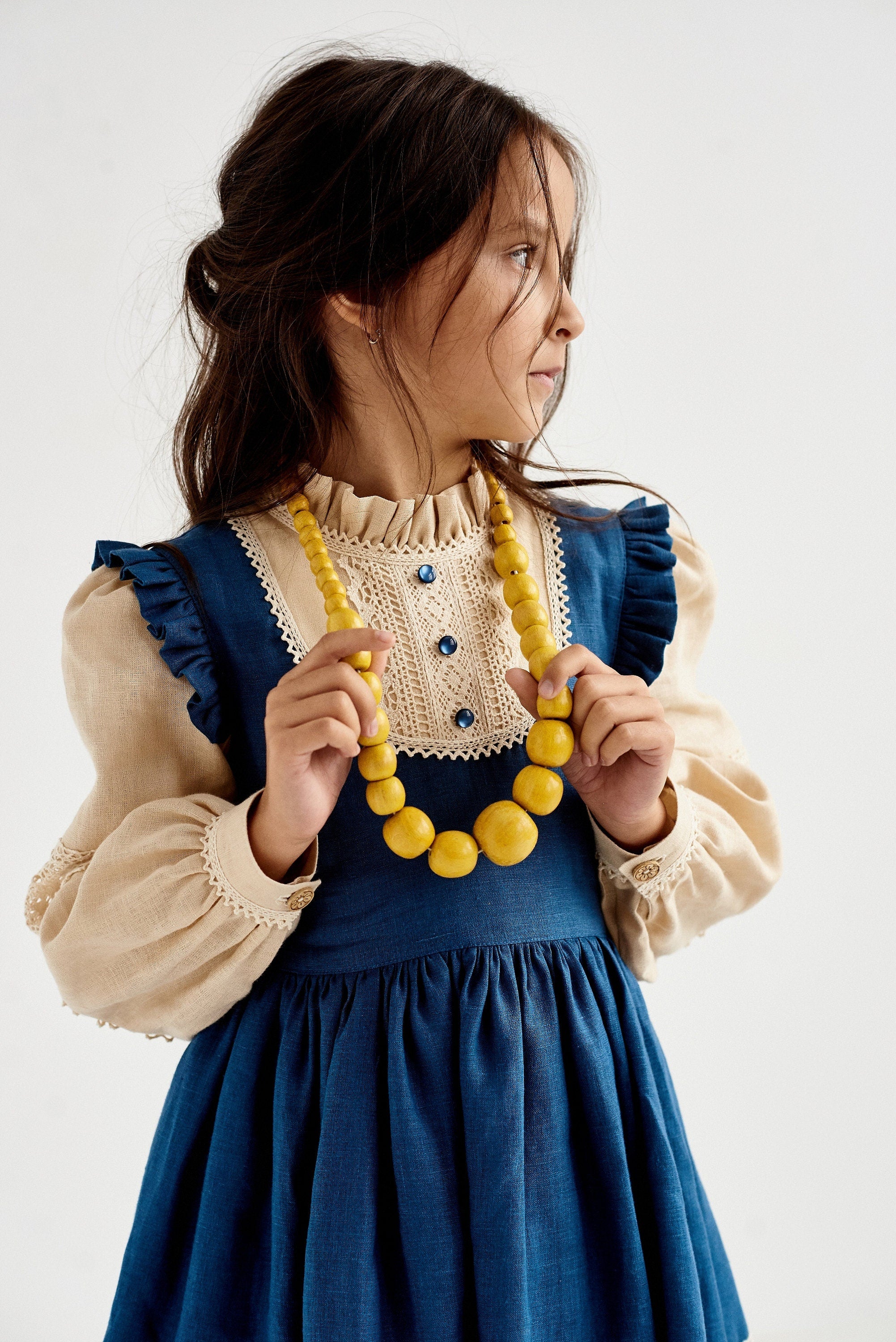 Young girl wearing a blue Ukrainian linen dress with puff sleeves, lace trim, and yellow bead necklace, in a traditional boho style.