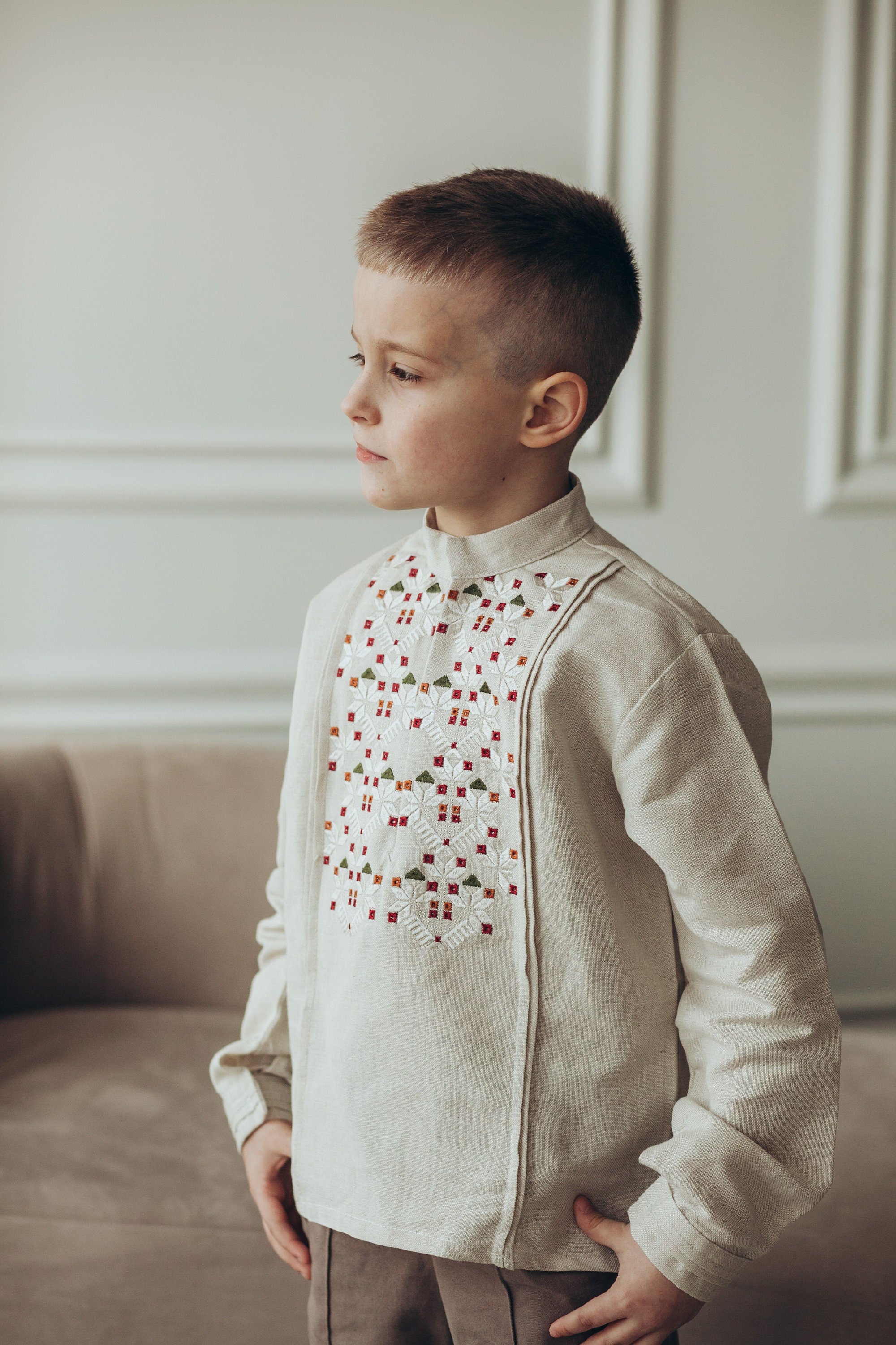 Young boy wearing a traditional Ukrainian embroidered vyshyvanka shirt in white linen with long sleeves, standing indoors.