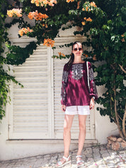 Woman wearing marsala boho Vyshyvanka blouse with white embroidery, standing against a leafy backdrop.