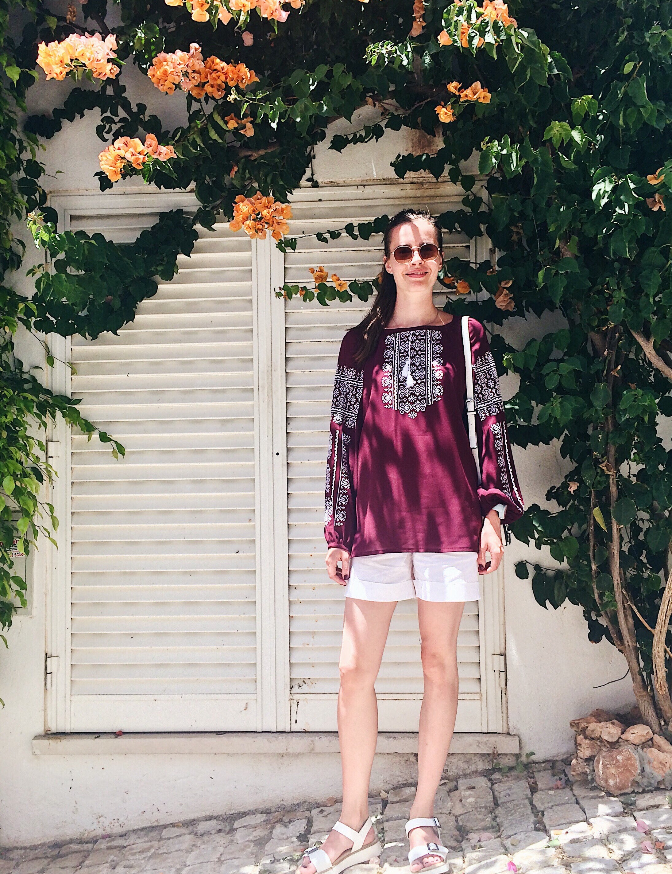 Woman wearing marsala boho Vyshyvanka blouse with white embroidery, standing against a leafy backdrop.