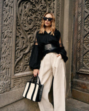 Woman wearing black embroidered blouse with tulle inserts, holding a stylish bag, stands against ornate wall. Boho ethnic chic fashion.