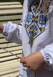Girl wearing Ukrainian Vyshyvanka dress with trident embroidery, holding yarn. Traditional boho toddler trapeze dress in natural cotton.