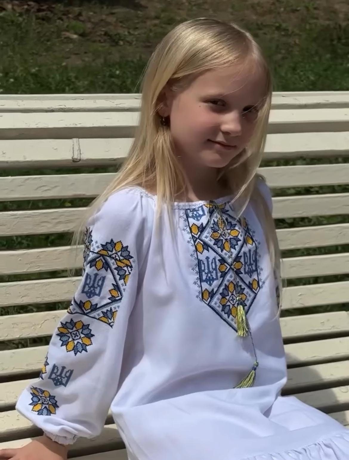 Girl wearing Ukrainian Vyshyvanka embroidered dress with Trident symbol, seated on bench, showcasing traditional boho style.