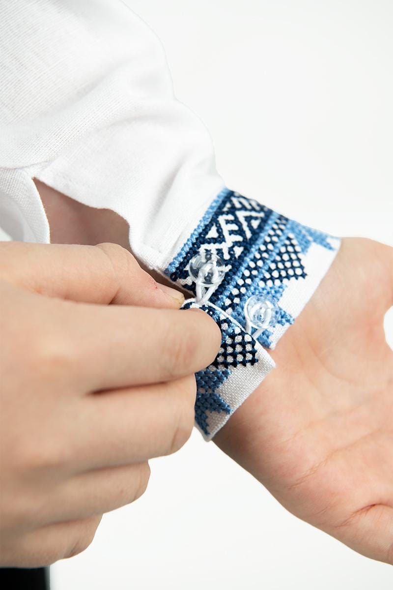 Close-up of a hand buttoning a Ukrainian white linen shirt with blue embroidery for boys, showcasing traditional patterns.