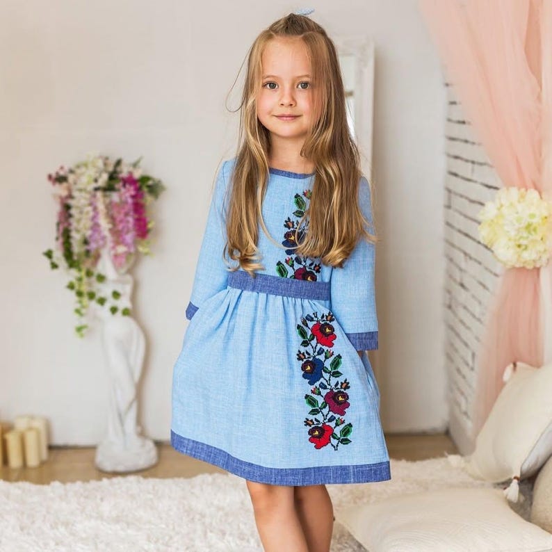 Girl wearing Ukrainian embroidered dress with floral vyshyvanka design, standing in decorated room with flowers.