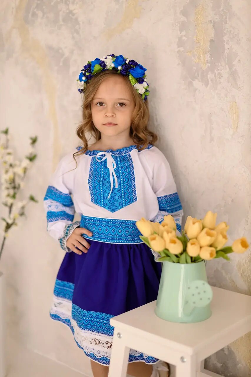 Girl in Ukrainian Hutsul folk costume with blue and lace details, wearing a floral headband, standing near flowers in a vase.