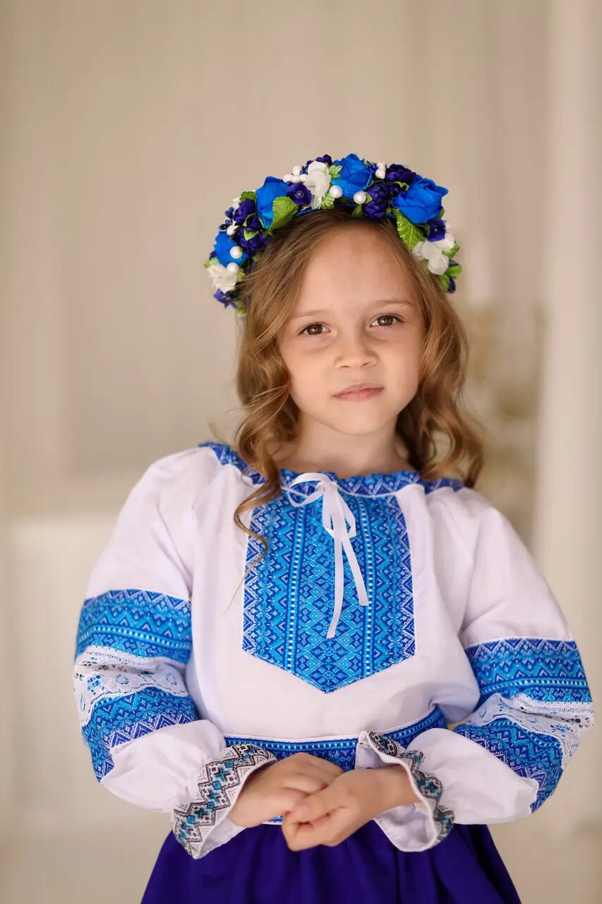 Girl wearing Ukrainian Hutsul folk costume with blue woven blouse, matching skirt and floral headband.