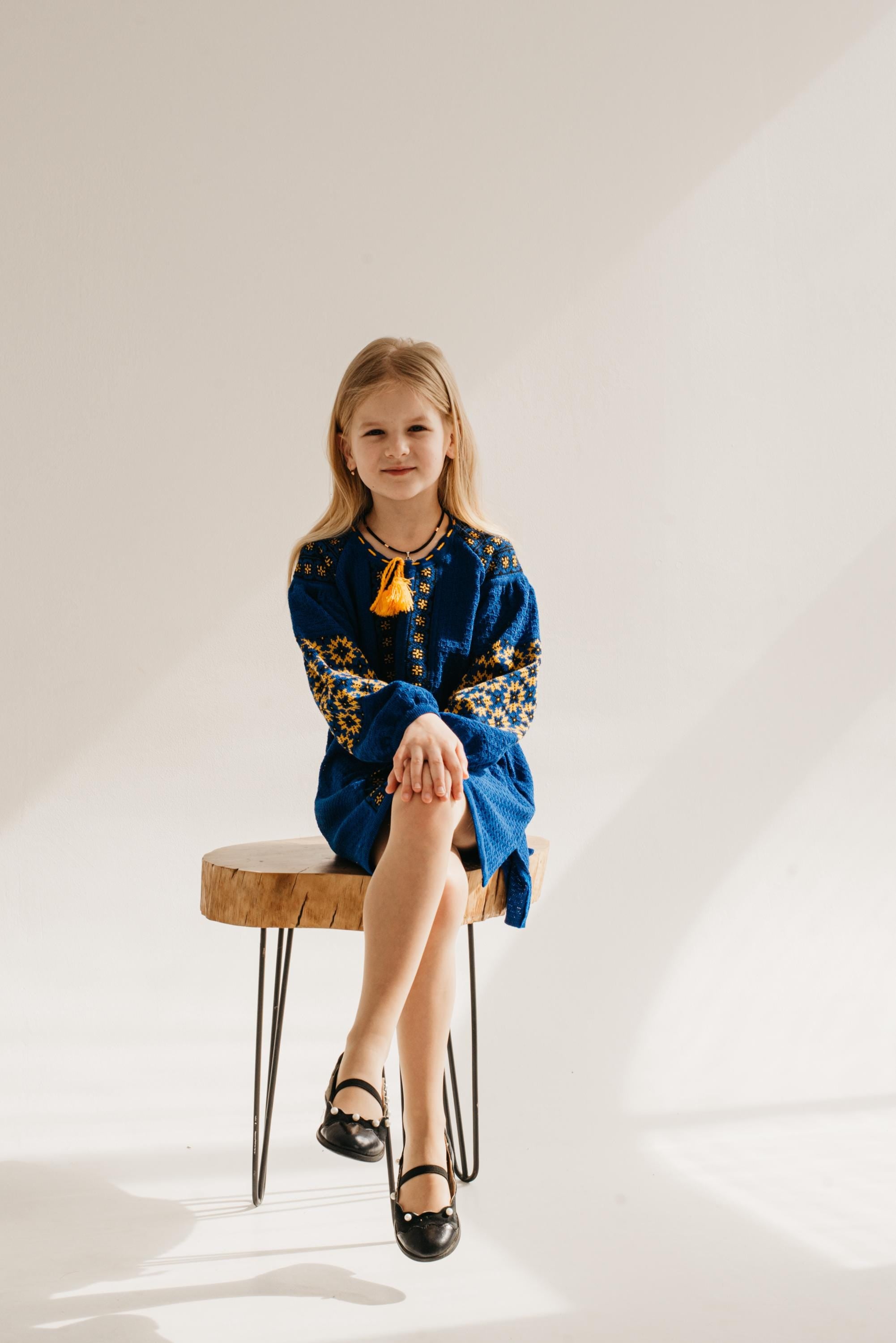 Young girl in a blue knitted dress with Ukrainian embroidery, sitting on a stool against a light background.