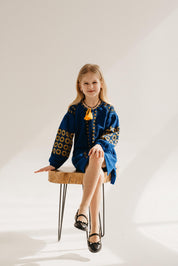 Young girl wearing a blue Ukrainian knitted dress with traditional yellow embroidery, sitting on a wooden stool.