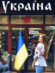 Woman wearing a white linen vyshyvanka blouse with red embroidery, standing in front of a Ukrainian shop entrance with flag.