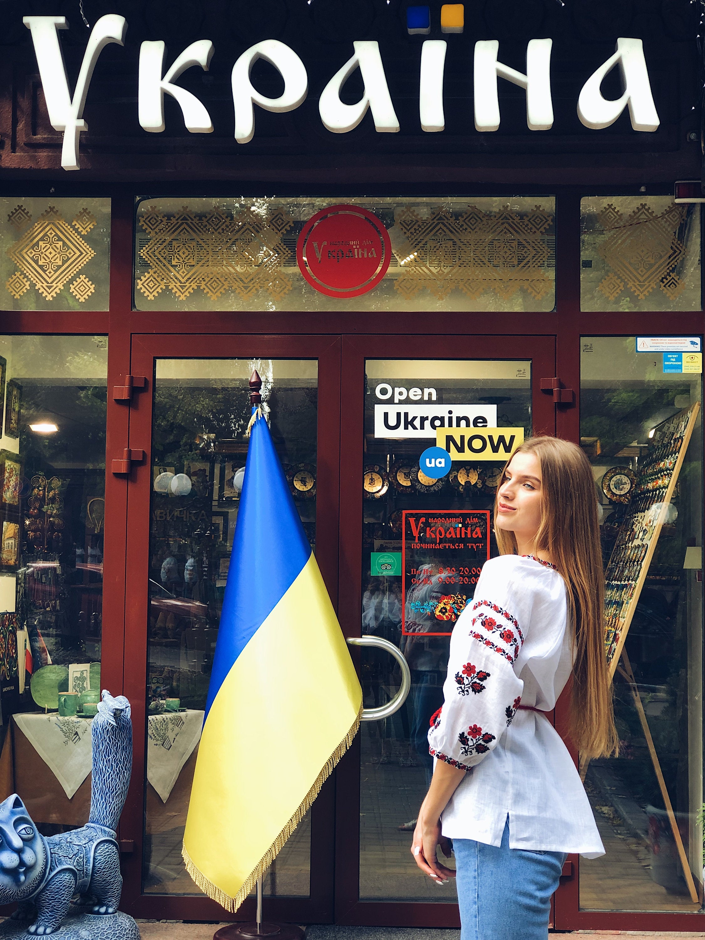 Woman wearing a white linen vyshyvanka blouse with red embroidery, standing in front of a Ukrainian shop entrance with flag.