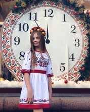 Girl in white linen embroidered dress stands in front of large clock, wearing a floral crown. Traditional Ukrainian Vyshyvanka outfit.