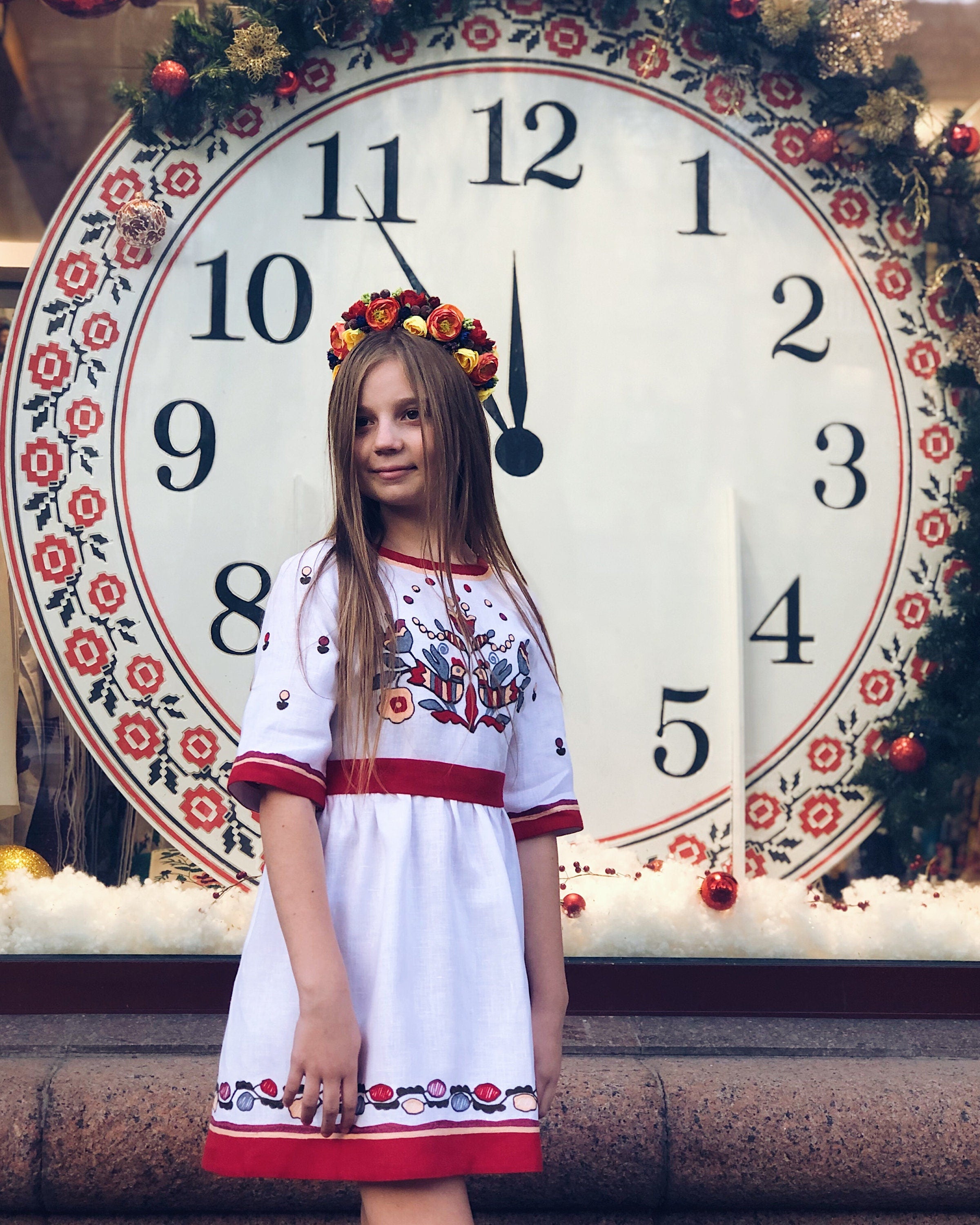 Girl in white linen embroidered dress stands in front of large clock, wearing a floral crown. Traditional Ukrainian Vyshyvanka outfit.