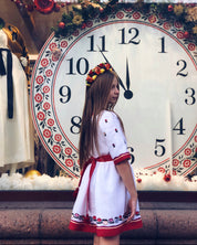 Girl wearing white embroidered dress standing in front of large decorative clock