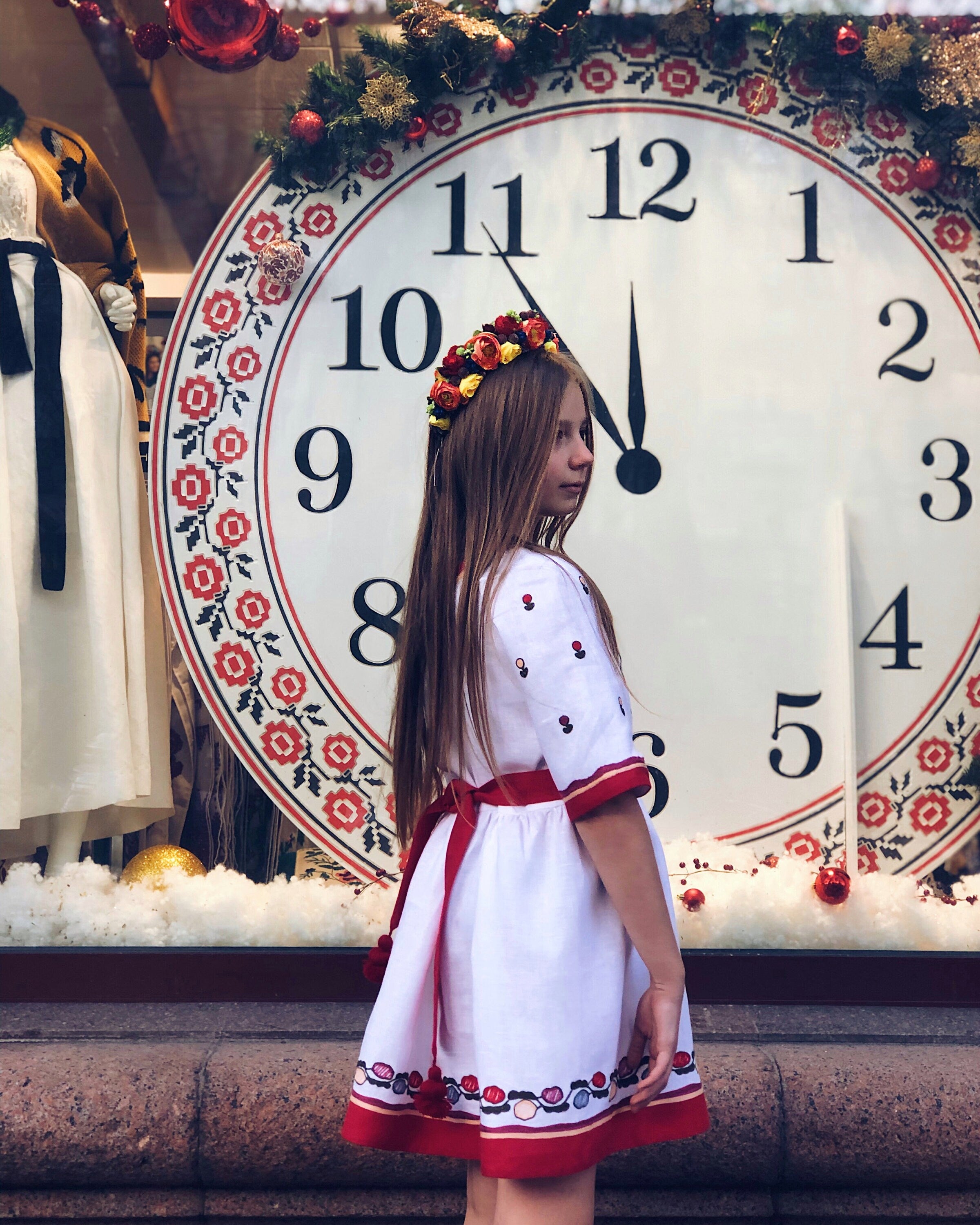Girl wearing white embroidered dress standing in front of large decorative clock