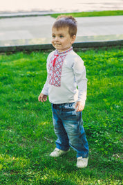 Boy wearing traditional Ukrainian vyshyvanka, embroidered white shirt for toddlers, in a grassy park setting.