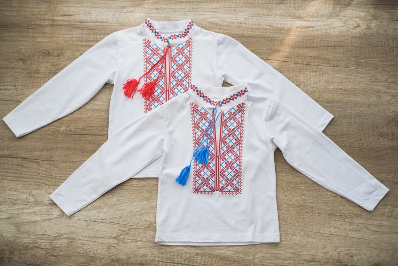 Traditional Ukrainian vyshyvanka embroidered shirts for babies and toddlers, featuring red and blue patterns, displayed on wooden surface.