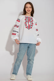 Child wearing Ukrainian white vyshyvanka shirt with red embroidery, blue jeans, standing, showcasing traditional kids' folk blouse.