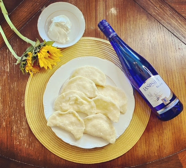 Plate of homemade Ukrainian varenyky dumplings with a side of sour cream, bottle of wine, and sunflowers on a wooden table.