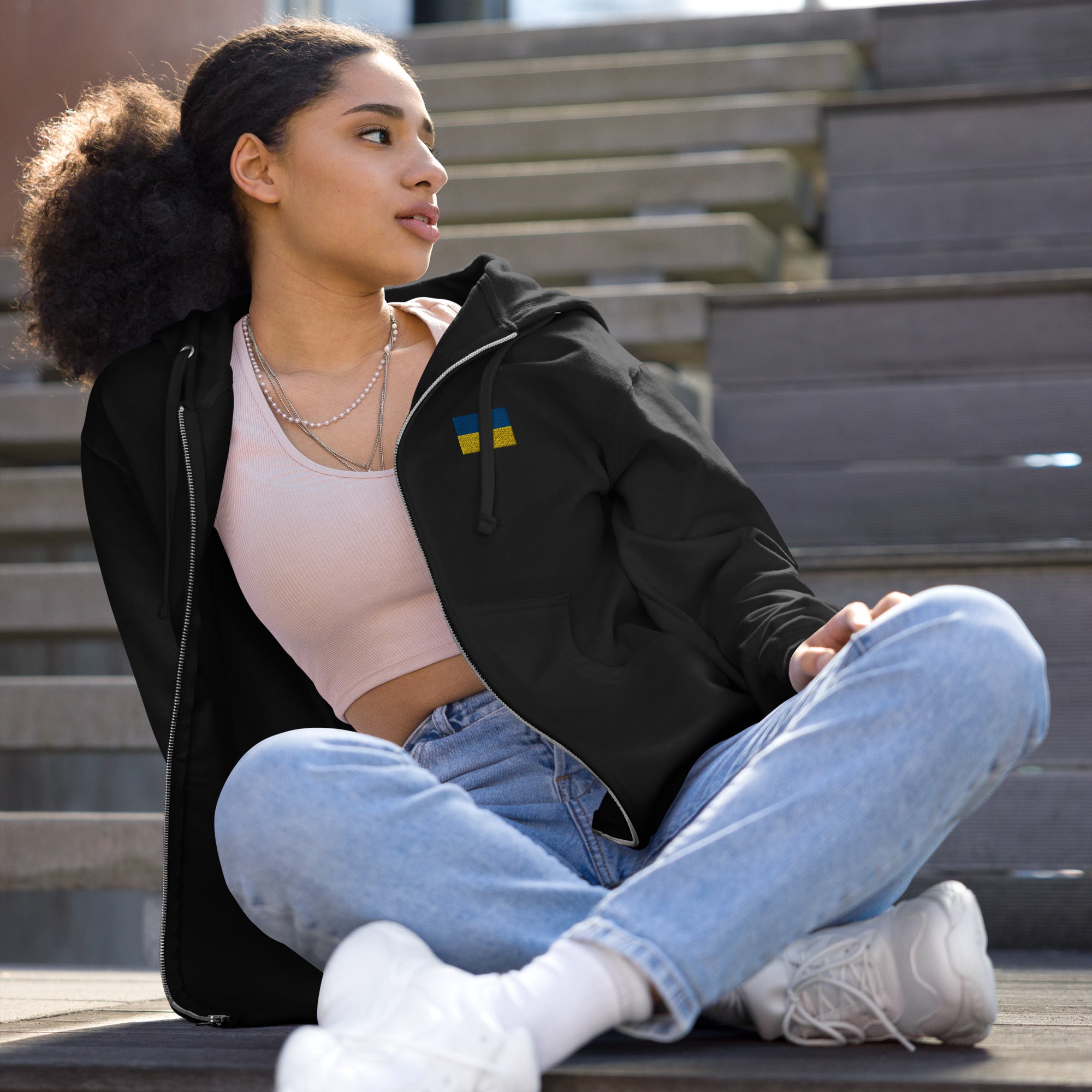 Woman wearing Ukrainian Flag fleece zip-up hoodie from Ukrainian Vibe Trizub Collection, sitting outdoors on steps.