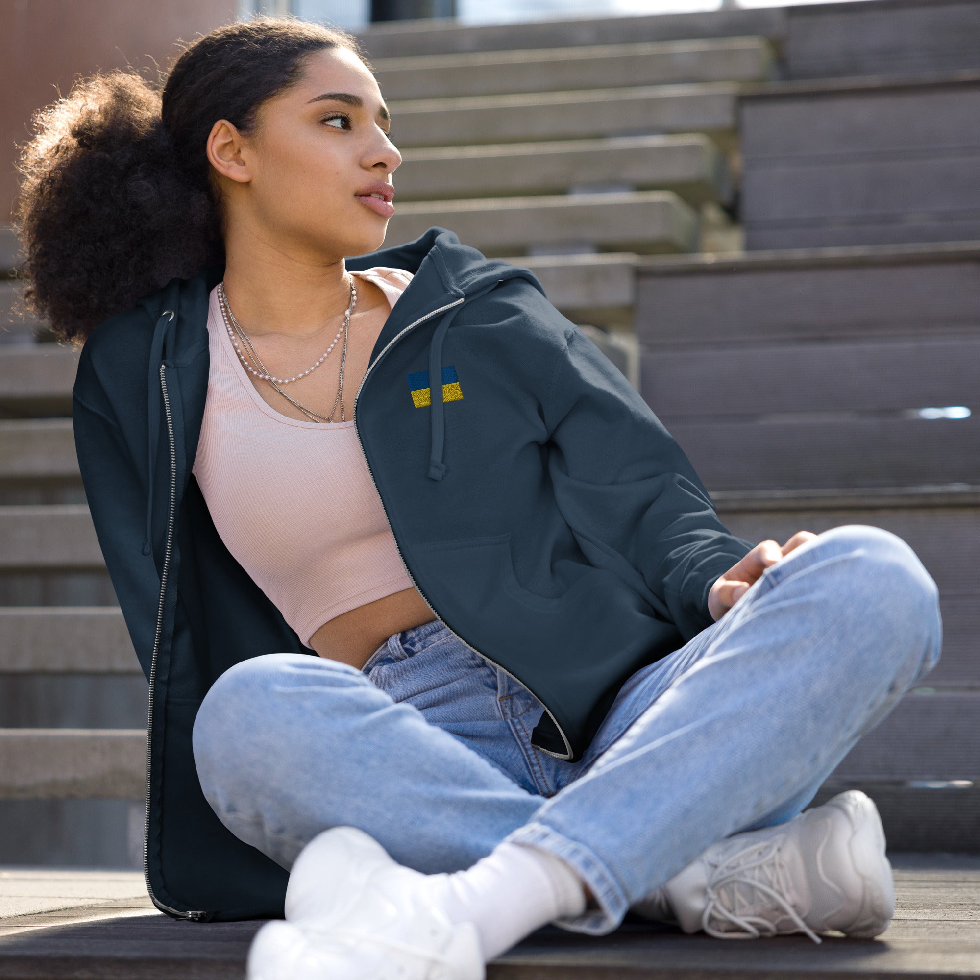 Woman wearing Ukrainian Flag fleece zip-up hoodie sitting on stairs, stylish casual outfit with jeans and sneakers.