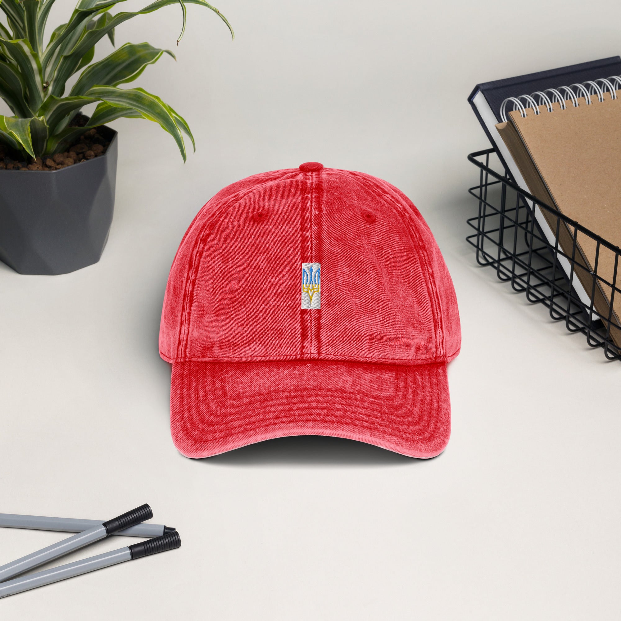 Red vintage cotton twill cap with embroidered Ukrainian Tryzub, featuring a washed-out effect and adjustable brass buckle, displayed on a desk.