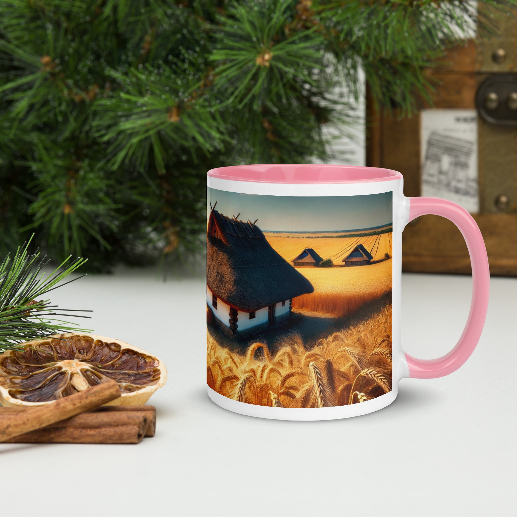 Ceramic mug with Ukrainian cottage design in wheat field, pink handle, on table with pine branches; 11 oz size shown.