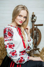 Woman wearing embroidered Ukrainian white sorochka blouse with red and black traditional patterns, sitting near spinning wheel.
