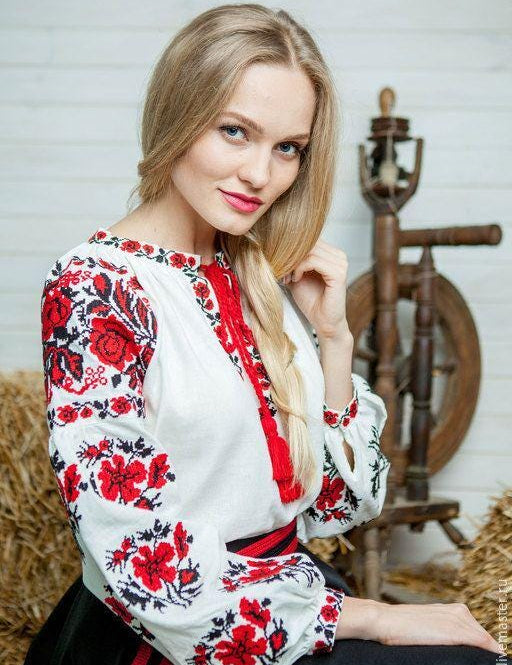 Woman wearing embroidered Ukrainian white sorochka blouse with red and black traditional patterns, sitting near spinning wheel.