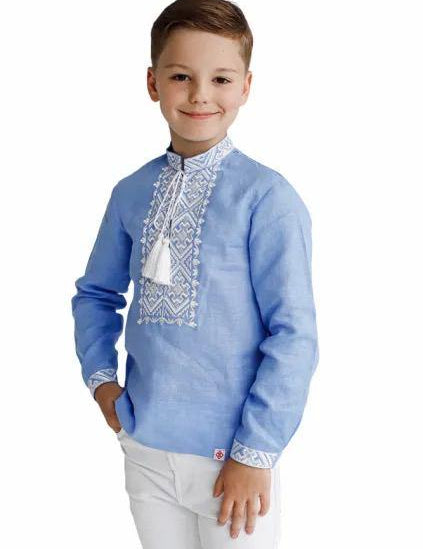 Boy wearing a blue linen embroidered Ukrainian Vyshyvanka with white cross-stitch, standing with a smile.