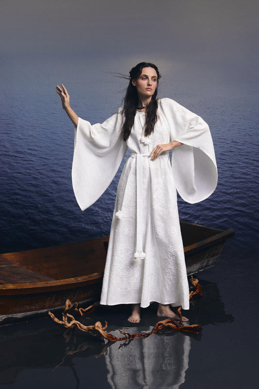 Woman in a white linen Ukrainian embroidered vyshyvanka wedding dress, standing by a boat on a calm lake.